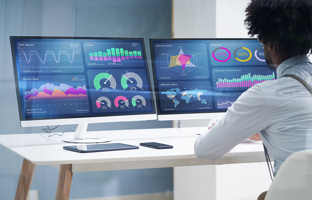Black business man sitting in front of two computer screens displaying colorful business data