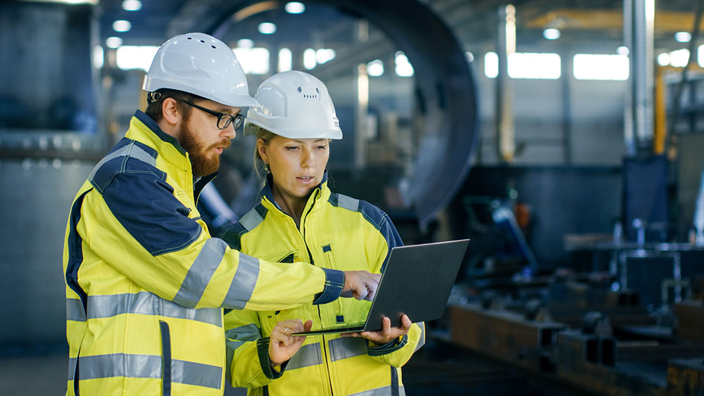 Male and female industrial engineers in hard hats discuss while using laptop Manufacturing plant