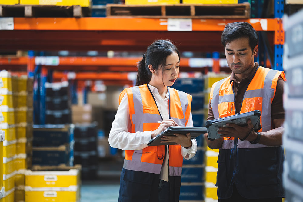 Female and male workers wearing reflective vests standing in a logistics warehouse while using digital tablets.