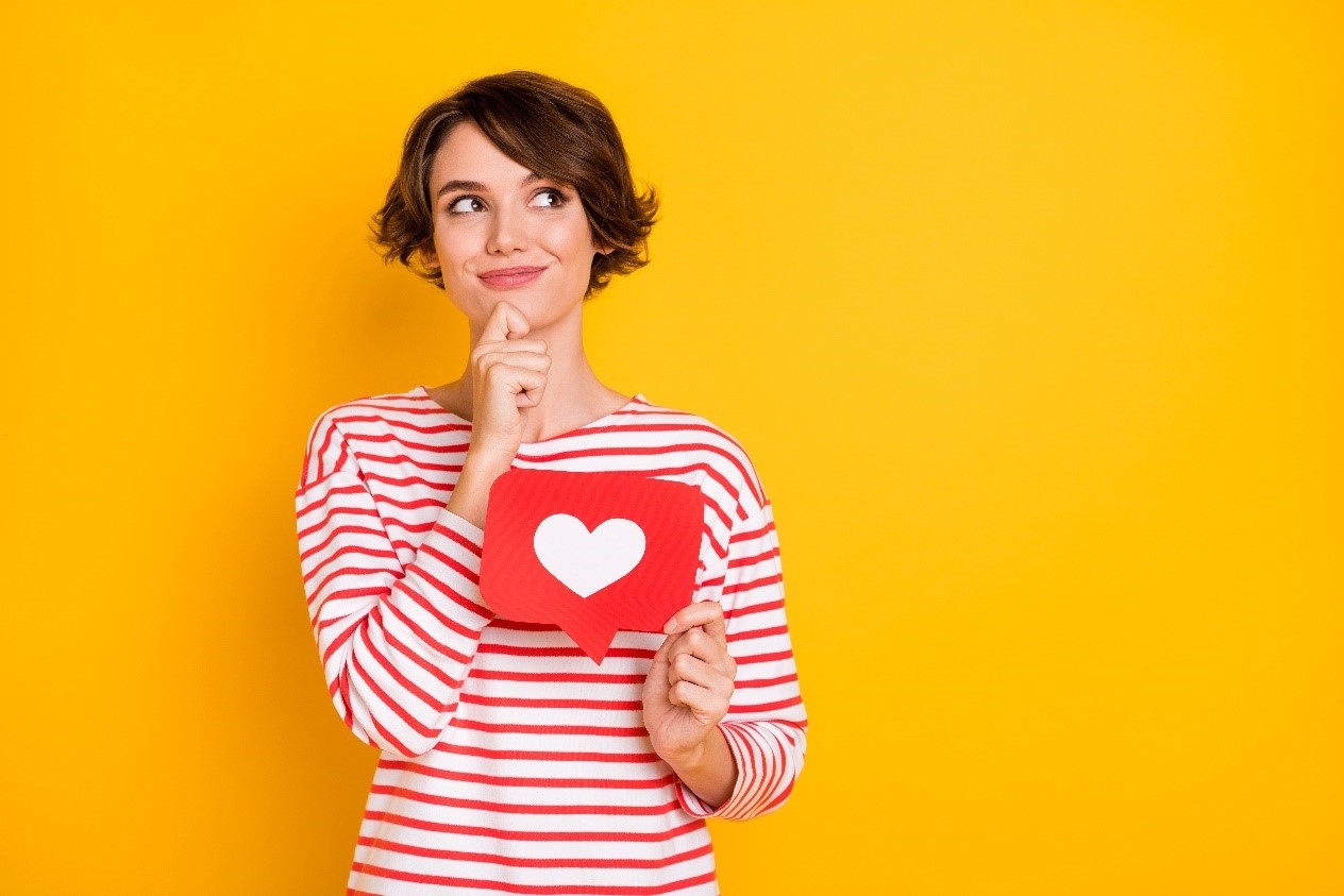 A young woman with short brown hair, wearing a red and white striped shirt, holds a red speech bubble with a white heart. She's thoughtfully touching her chin, gazing off to the side against a bright yellow backdrop.