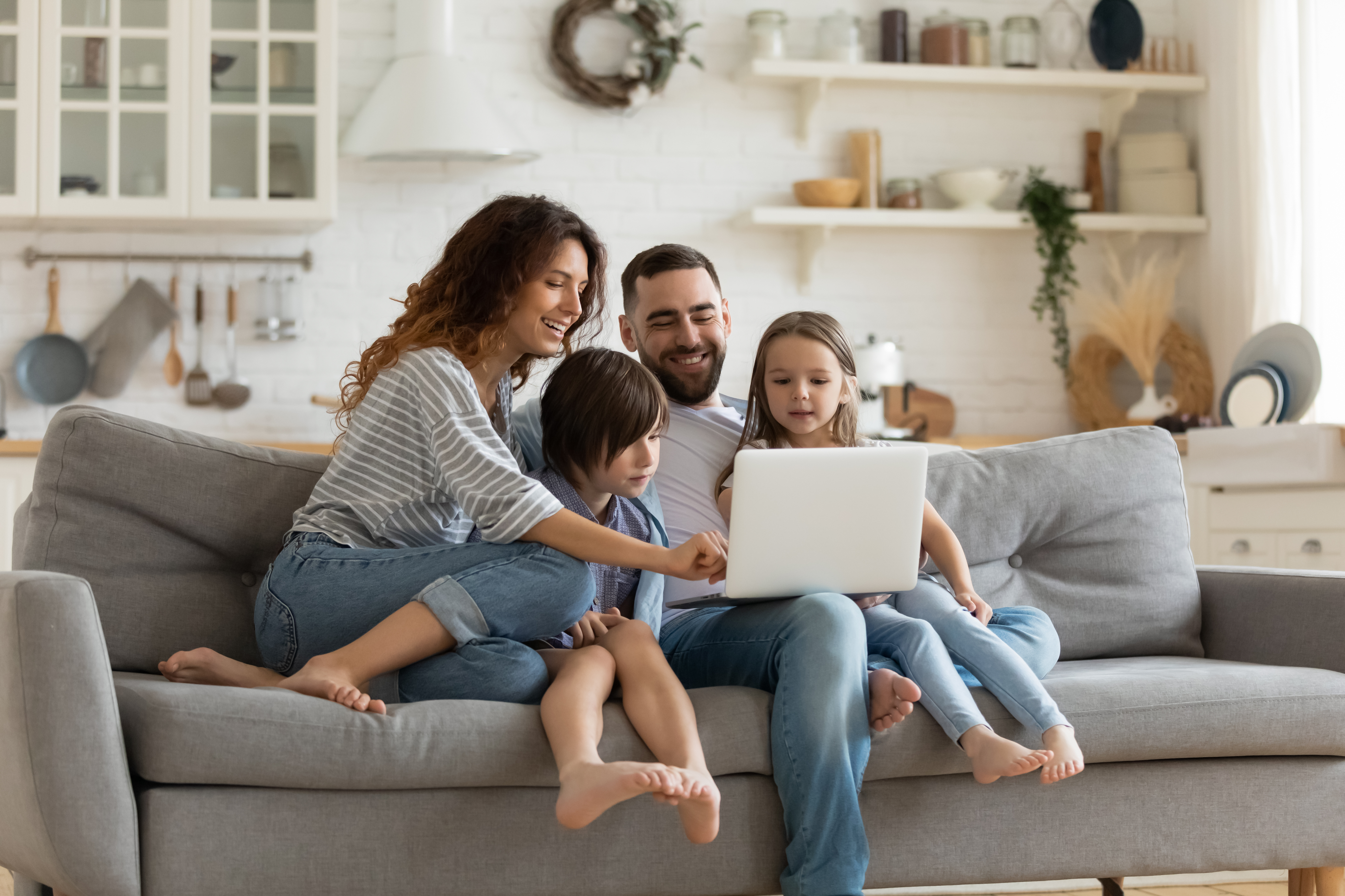 A happy family of four — a mother, father, son, and daughter — sitting together on a gray couch in their living room, all looking at a laptop computer that rests on the father's lap.