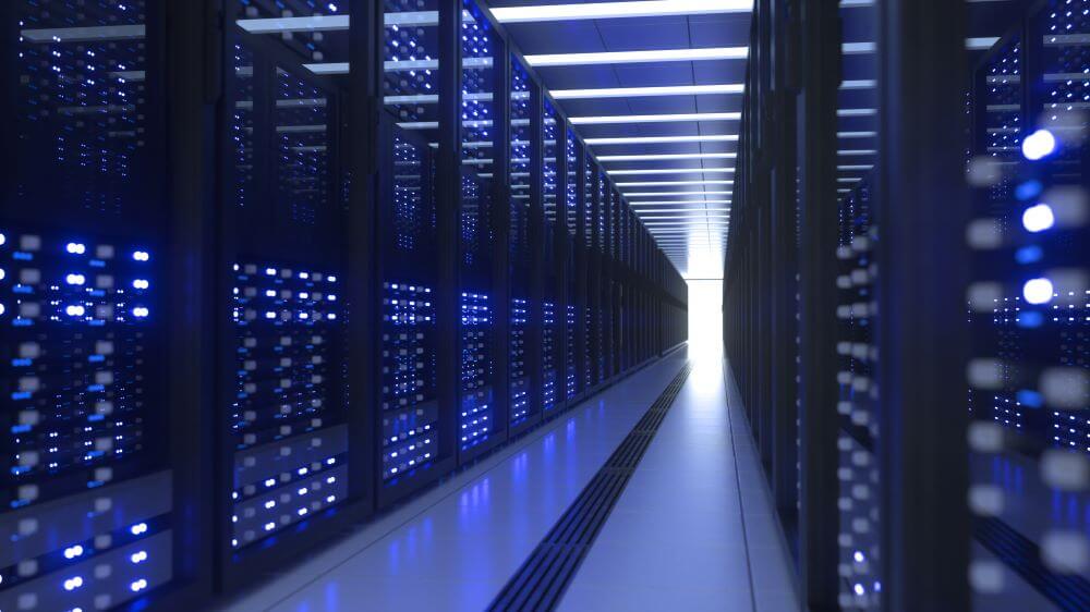 A long corridor of server racks in a modern data center. The servers are lit with blue lights, and the hallway extends to a bright light at the end.