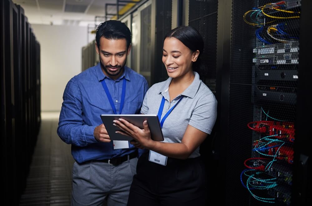 A man and a woman, both wearing company ID badges, collaborate in a server room. They are reviewing data on a tablet computer, surrounded by racks of network equipment and cables.