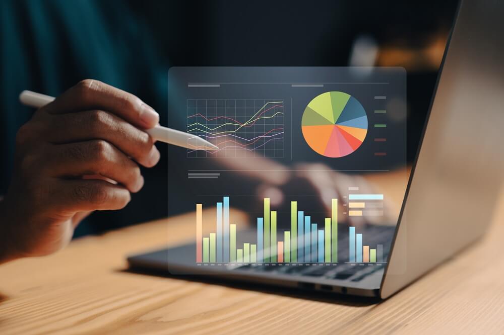 Close-up of a hand using a stylus to interact with a holographic display of financial data on a laptop. The display shows line graphs, bar charts, and a pie chart.