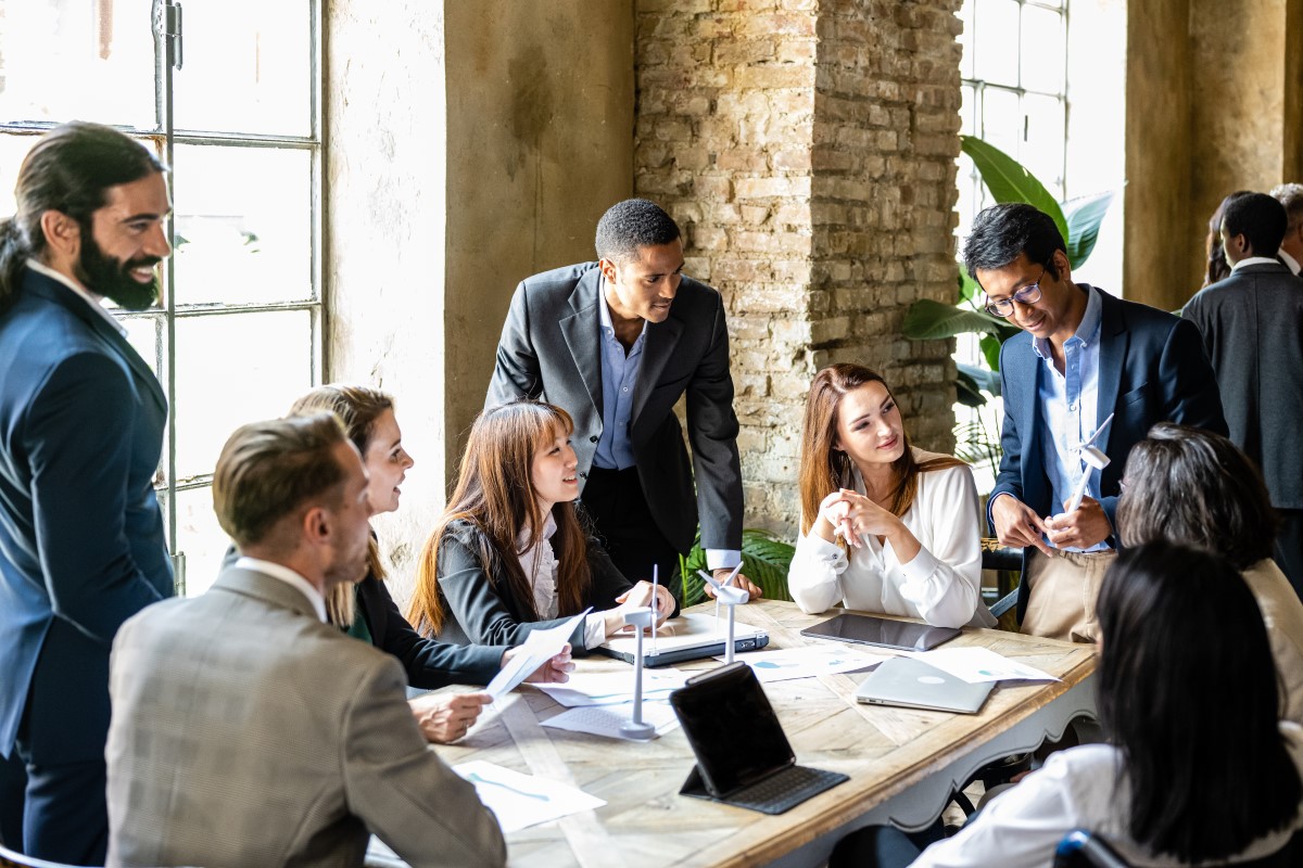 A diverse group of five people gathered around a wooden table in an office. A whiteboard with a bar graph drawn on it is visible in the background, suggesting a business or project-related meeting. They are likely discussing business strategies, reviewing data, or planning a project