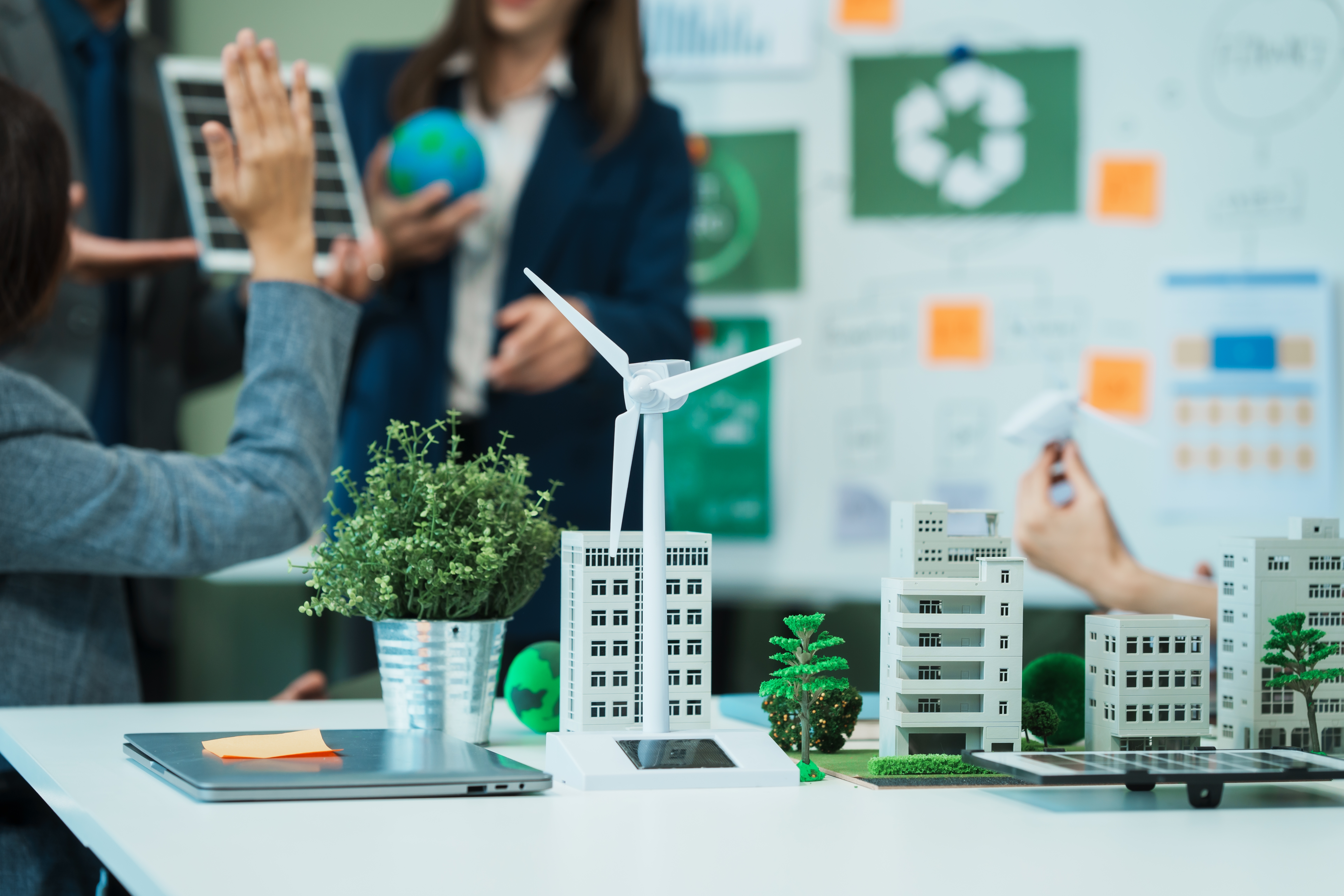 A tabletop 3D model of a sustainable city, featuring miniature buildings, a white wind turbine, and greenery, with a solar panel array in the foreground. In the blurry background, people are collaborating around a whiteboard with environmental symbols and a person holds a miniature globe, indicating a discussion about green energy and eco-friendly urban planning.